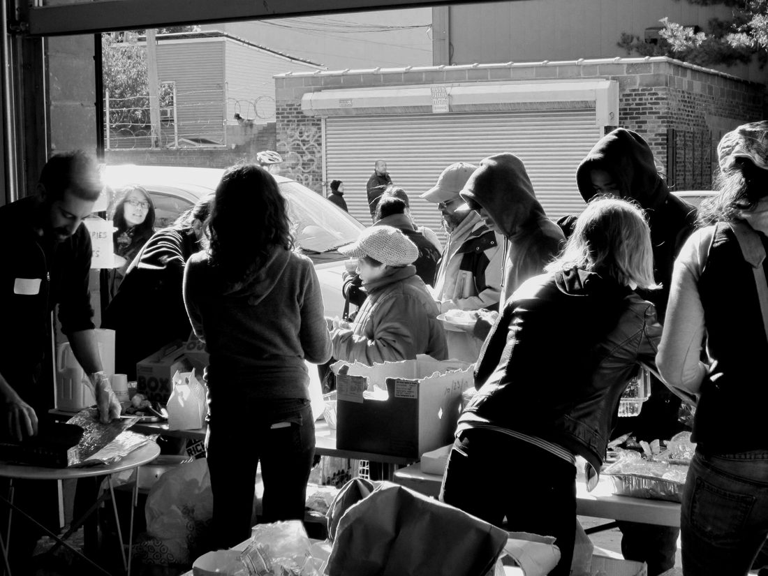 Black-and-white photograph of a Recovers relief distribution: volunteers handing out food and supplies from tables in a garage bay, neighbours queuing with plates.