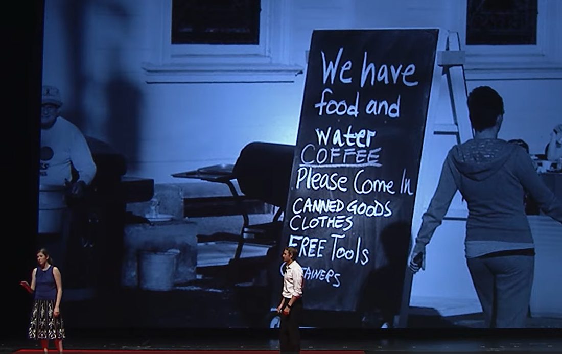 Caitria O'Neill presenting the Recovers work on stage; projected behind her is the Monson relief centre's hand-lettered sign: we have food and water, coffee, please come in, canned goods, clothes, free tools.