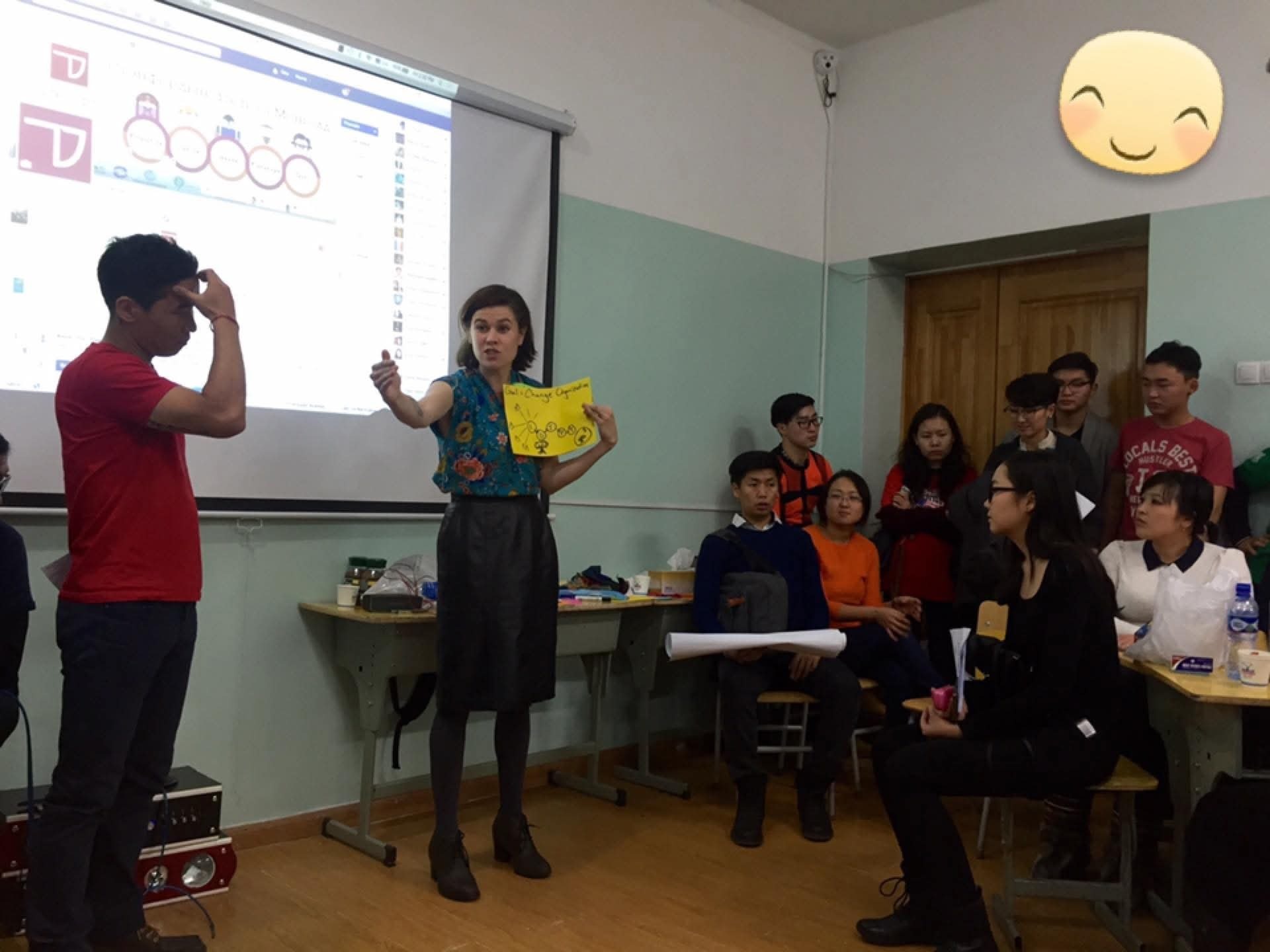 Caitria presenting at a workshop: standing at the front of a classroom of seated participants, a projector screen behind her, holding up a hand-drawn yellow sheet of paper as she speaks.